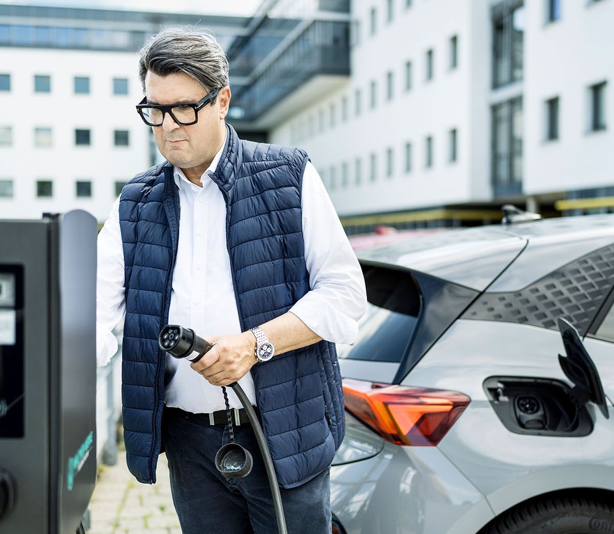 Mann mit AC-Ladestecker in der Hand an Ladesäule, dahinter ein E-Auto (Cupra)