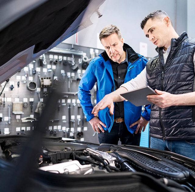 In der Auto-Werkstatt: Kunde und Kfz-Mechaniker bzw. Kfz-Mechatroniker stehen vor geöffneter Motorhaube. Der Kunde erhält eine Empfehlung, was repariert werden sollte, um den Verkaufspreis des Gebrauchtwagens zu erhöhen.