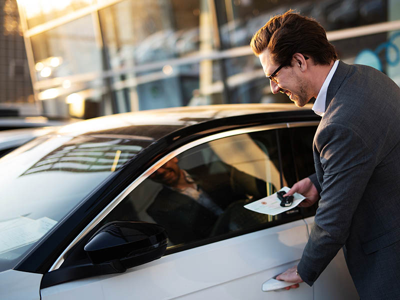 Mann mit Fahrzeugschein in der Hand sowie Autoschlüssel an der Autotür seines neu gekauften Pkw